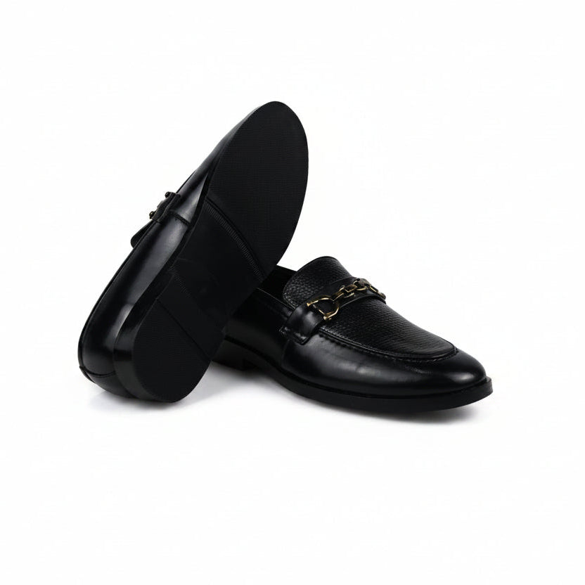 A pair of black leather loafers with a gold-tone chain detail across the vamp, displayed on a white fabric backdrop.