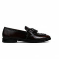 A single dark brown leather loafer with a decorative tassel strap across the vamp, shown in profile against a wrinkled white studio backdrop.