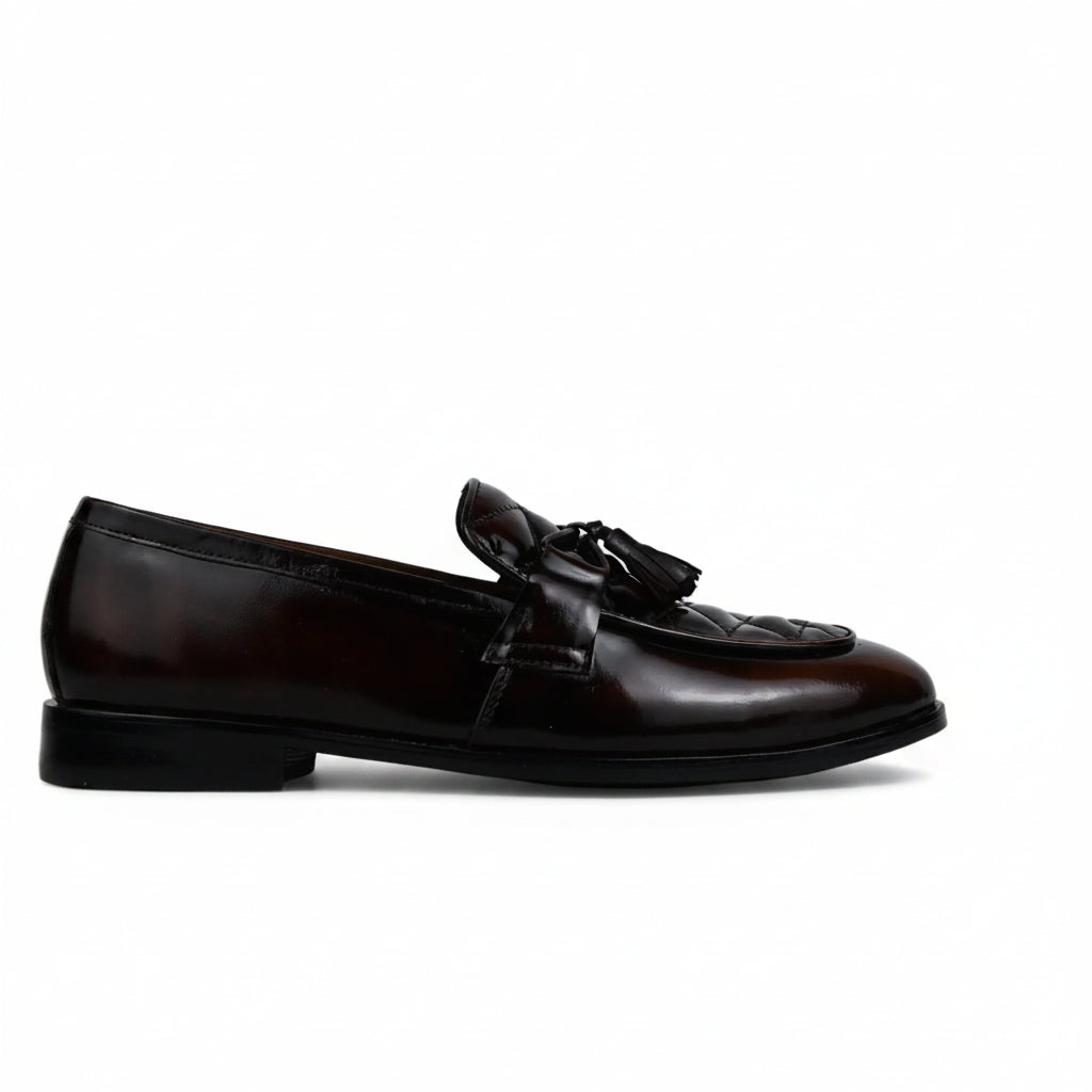 A single dark brown leather loafer with a decorative tassel strap across the vamp, shown in profile against a wrinkled white studio backdrop.