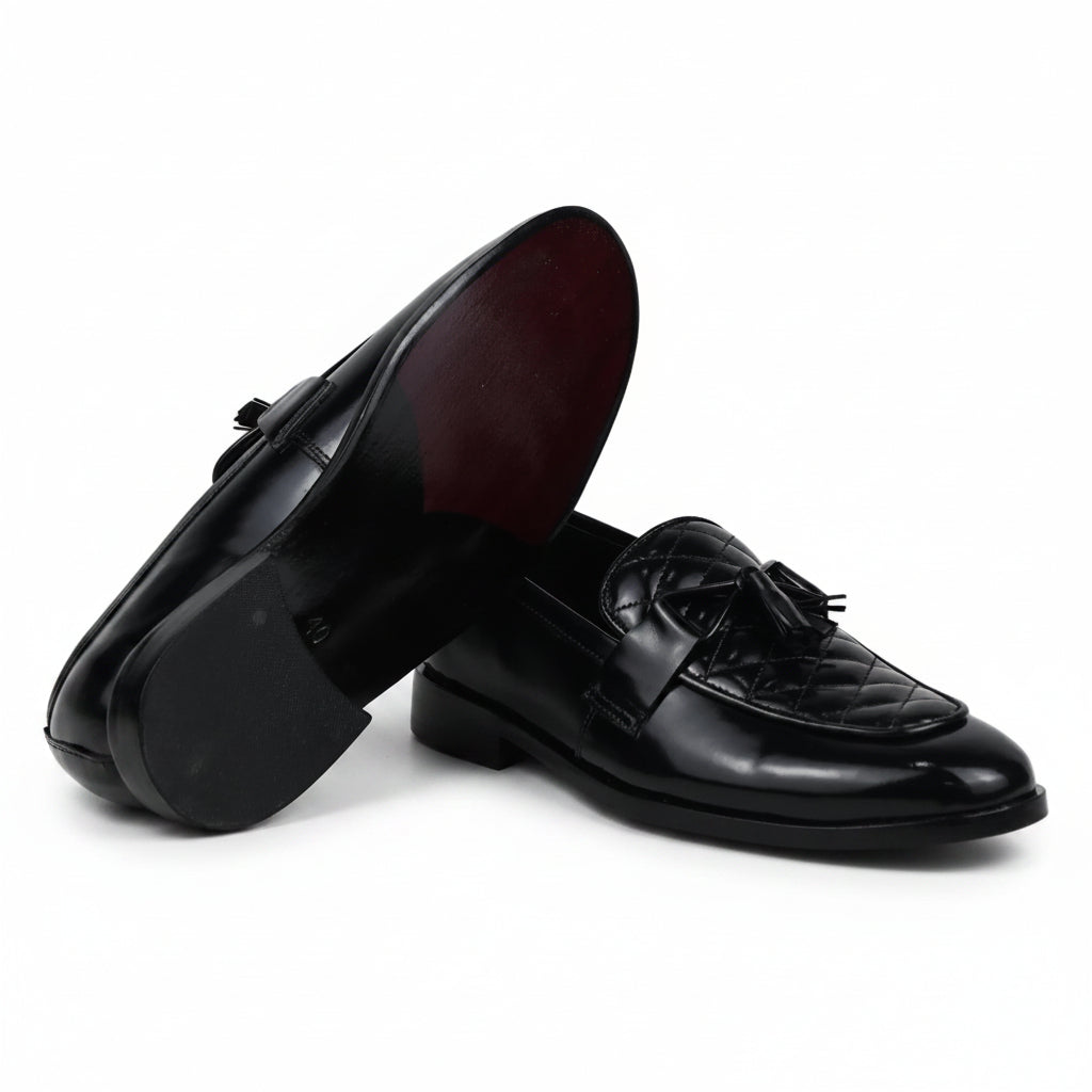 A pair of black leather penny loafers, with one shoe standing upright to show the dark red underside and the other lying flat to display the quilted vamp and tassel detail, set against a white studio backdrop.
