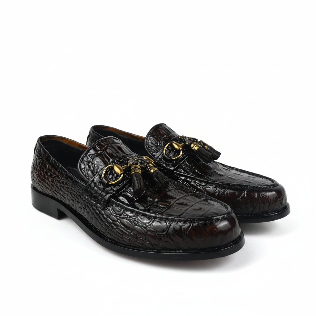 A pair of dark brown crocodile-embossed leather loafers with gold-tone horsebit details and tassel straps, displayed on a white studio background.