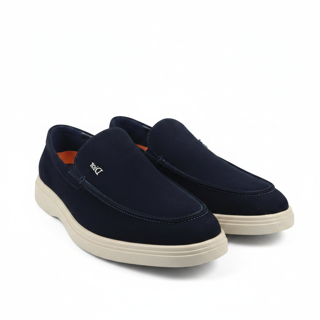 A pair of Dior slip-on loafers in navy blue suede with beige soles, displayed on a white studio background.