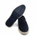 A pair of navy blue slip-on shoes with beige soles, shown with one shoe turned upside down to display the textured outsole, set against a plain white background.