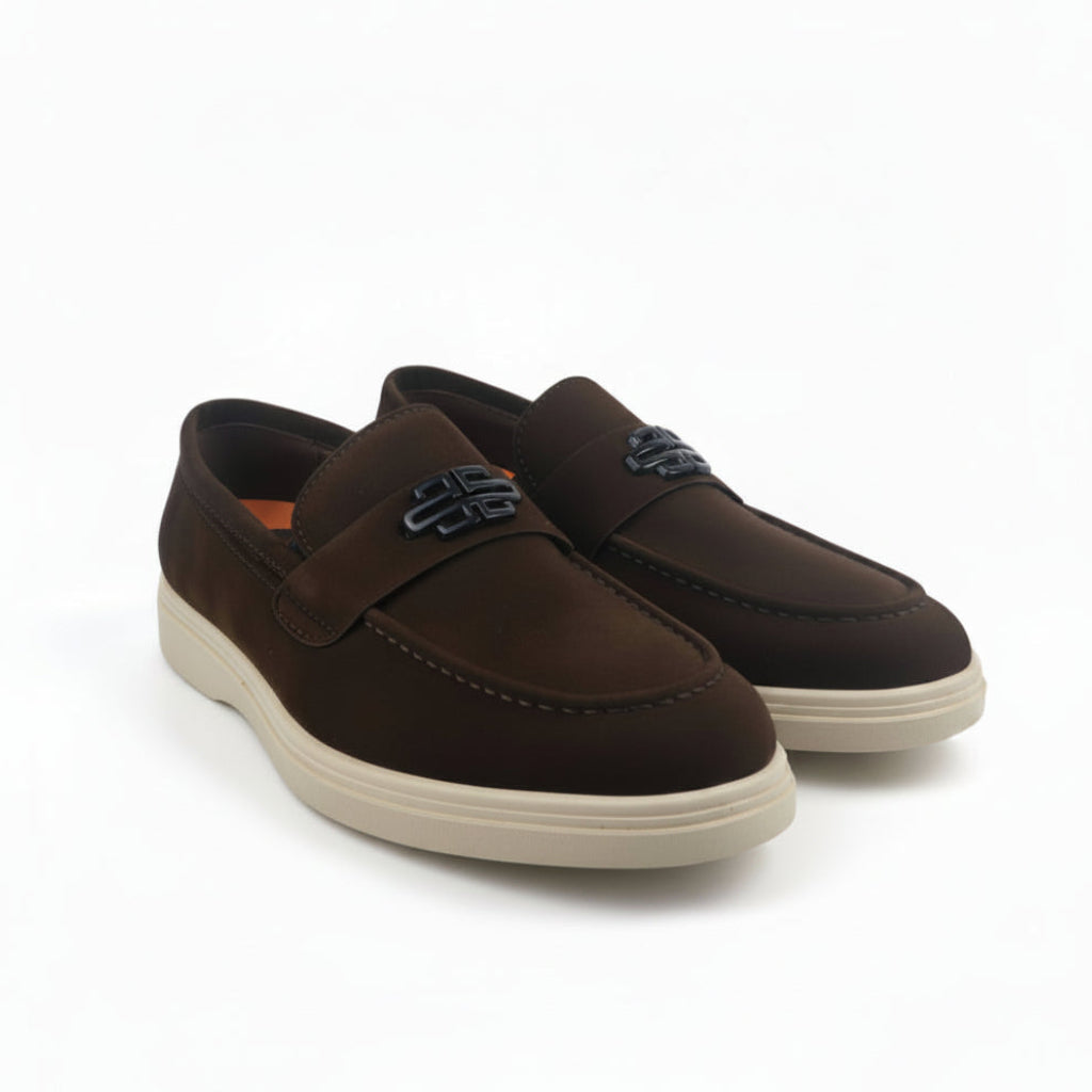 A pair of men's brown suede slip-on loafers with a decorative metal logo detail on the vamp and thick cream-colored soles, displayed against a white studio background.