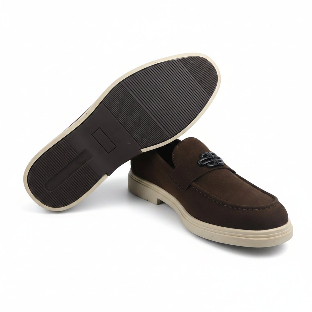A pair of dark brown suede loafers with cream-colored soles, with one shoe turned to show the textured black outsole. A small black metal logo is visible on the side of the shoe.