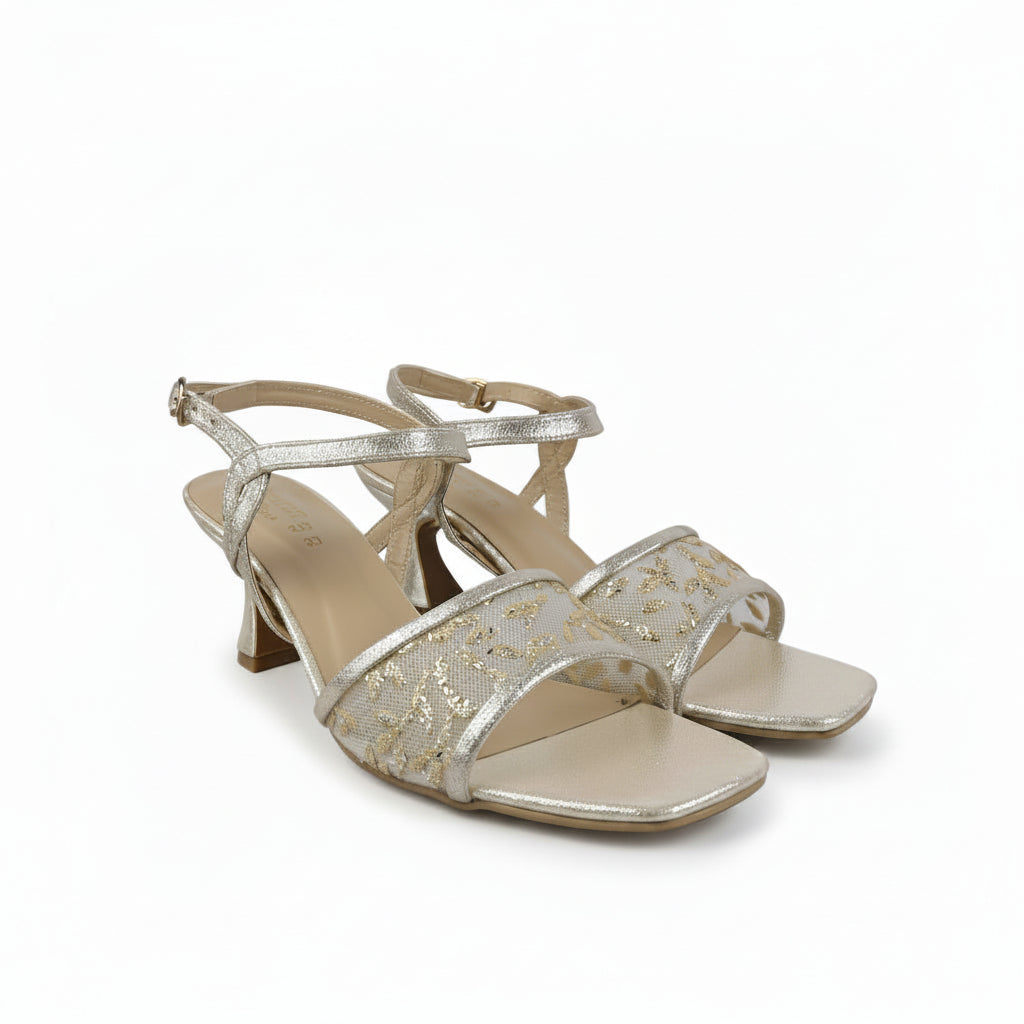 A pair of women's heeled sandals in a metallic champagne gold color, featuring an open toe, a square toe cap, and a thin ankle strap with a buckle closure. The straps are embellished with a textured, glittery pattern, and the insole is a light beige nude color.