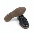 A black woven leather loafer shown from the side with its sole propped up, revealing a brown outsole. A light pink shoebox is partially visible on the right against a white background.