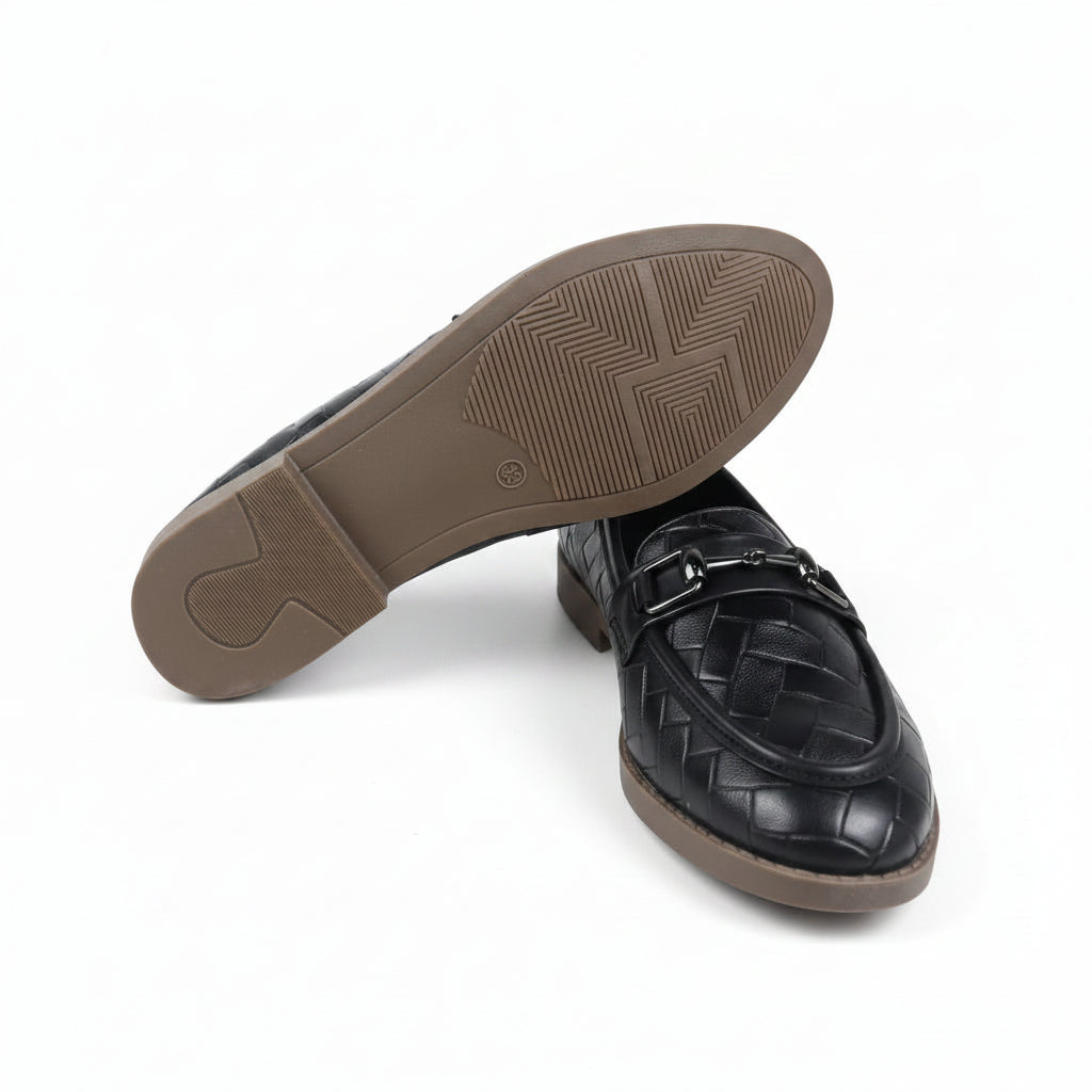 A black woven leather loafer shown from the side with its sole propped up, revealing a brown outsole. A light pink shoebox is partially visible on the right against a white background.