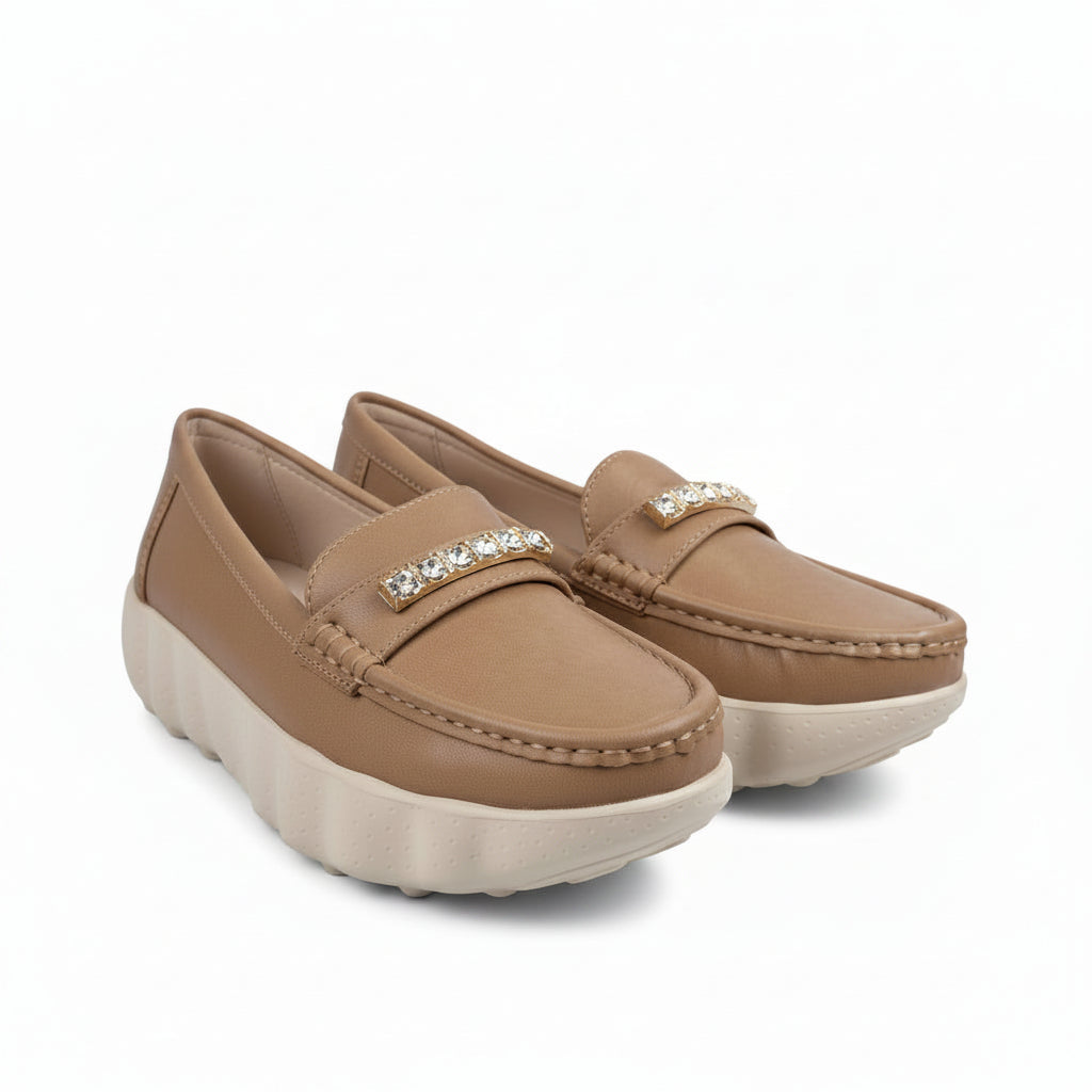 A pair of tan slip-on loafers with a decorative gold-tone embellished strap and chunky off-white platform soles, photographed on a white studio floor.