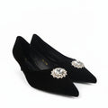 A pair of black pointed-toe kitten heels with jeweled floral embellishments on the toes, photographed on a light gray studio background.