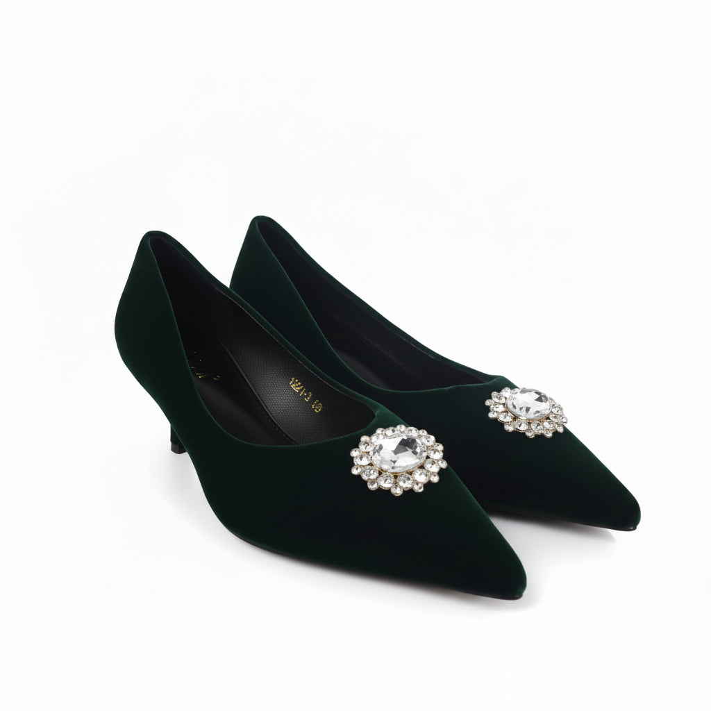 A pair of dark forest green velvet pointed-toe pumps with jeweled crystal brooch embellishments on the toes, displayed on a light gray studio floor.