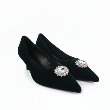 A pair of dark forest green velvet pointed-toe pumps with jeweled crystal brooch embellishments on the toes, displayed on a light gray studio floor.