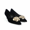 A pair of black pointed-toe kitten heels with textured gold flower embellishments on the toes, photographed on a light gray studio surface.