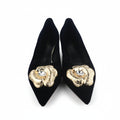 A pair of black pointed-toe high heels with large gold flower embellishments on the toes, photographed on a light gray surface with a shoe box partially visible on the right.
