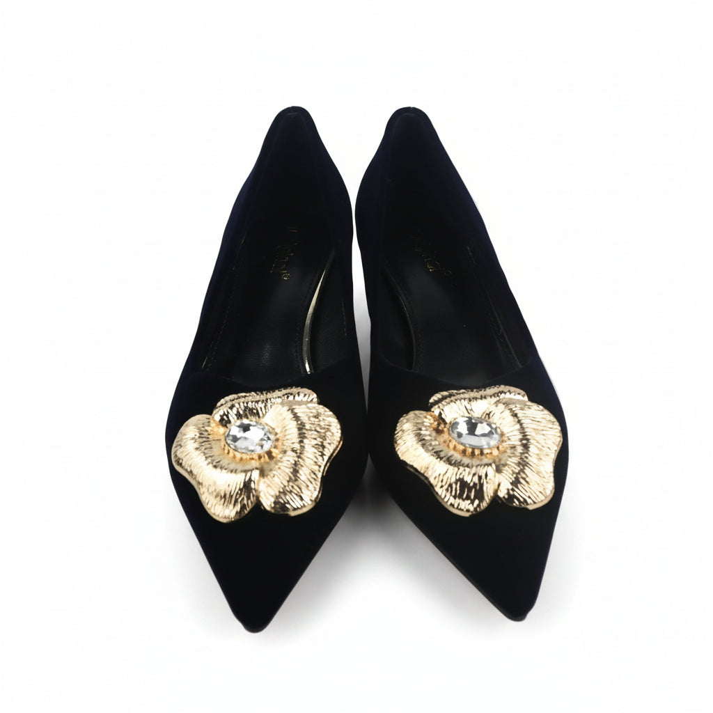 A pair of black pointed-toe high heels with large gold flower embellishments on the toes, photographed on a light gray surface with a shoe box partially visible on the right.