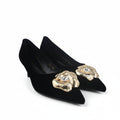 A pair of black pointed-toe kitten heels with ornate gold lion-head embellishments on the toes, photographed on a light gray studio surface.