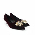 A pair of pointed-toe kitten heels in deep burgundy velvet, each decorated with a large gold-tone floral embellishment on the toe area featuring a clear rhinestone center, shown on a white studio floor.