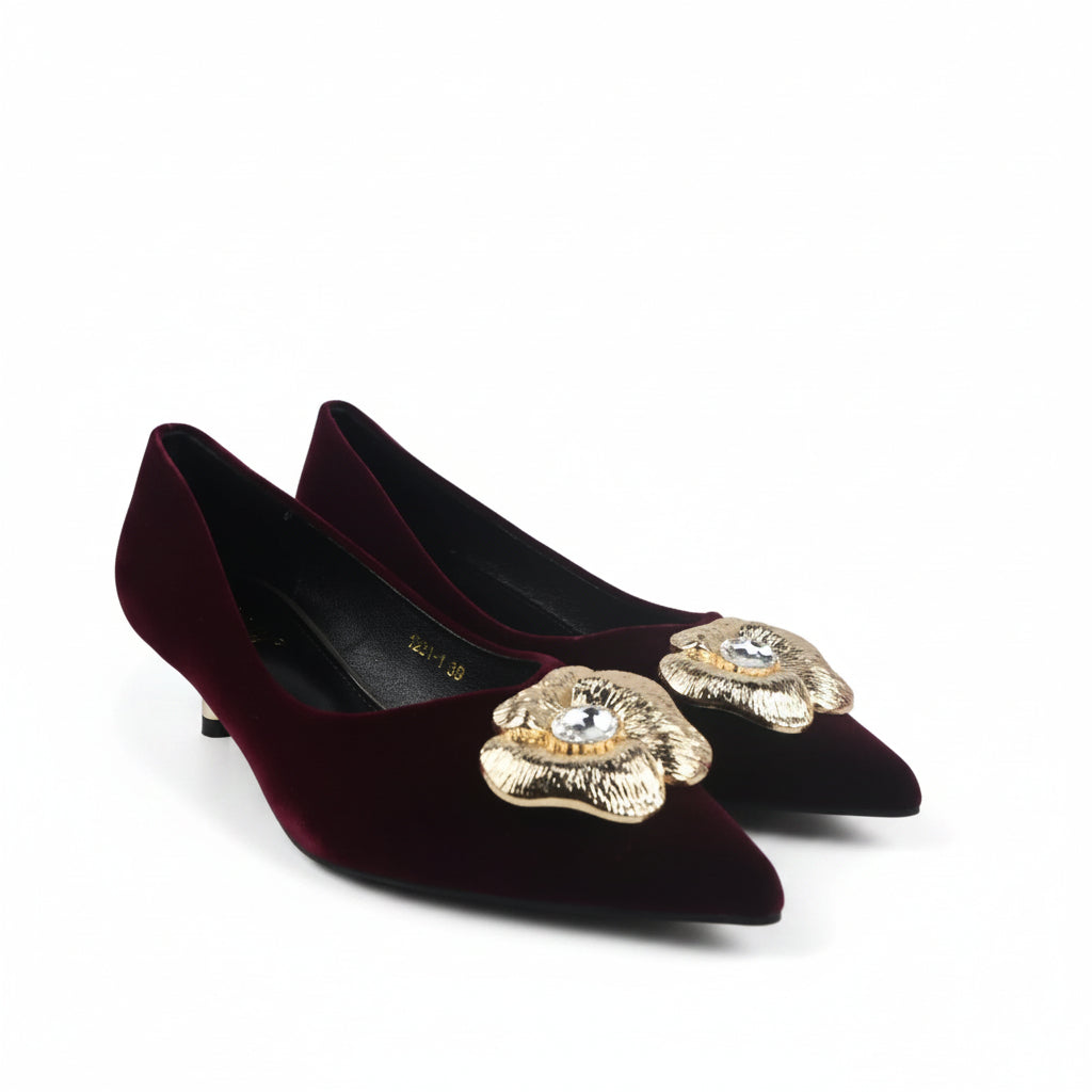A pair of pointed-toe kitten heels in deep burgundy velvet, each decorated with a large gold-tone floral embellishment on the toe area featuring a clear rhinestone center, shown on a white studio floor.