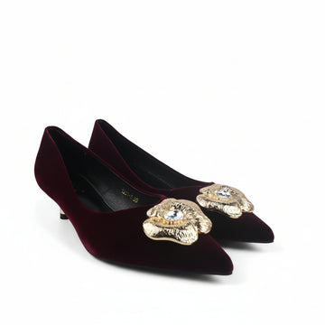 A pair of pointed-toe kitten heels in deep burgundy velvet, each decorated with a large gold-tone floral embellishment on the toe area featuring a clear rhinestone center, shown on a white studio floor.