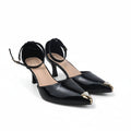 A pair of black patent leather high-heeled pumps with ankle straps and gold-tone toe caps, displayed on a light gray studio floor.