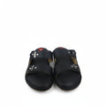 A pair of black leather slide sandals with double straps and silver metal rivets, shown next to a purple shoebox on a white background.
