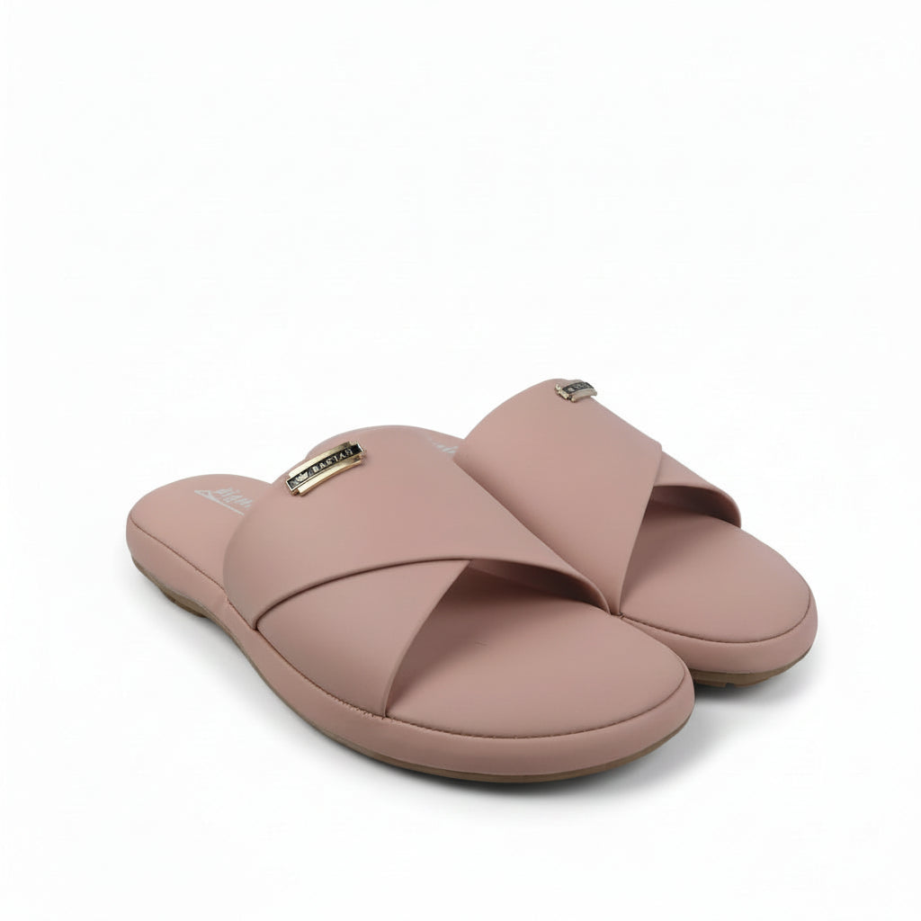 A pair of women's slide sandals in a dusty pink color, featuring a small gold-tone metal logo plaque on the strap, set against a white studio backdrop.