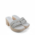 A pair of women's platform mule sandals with beige heels and white uppers, featuring multiple horizontal straps embellished with silver rhinestones, set against a white fabric backdrop.