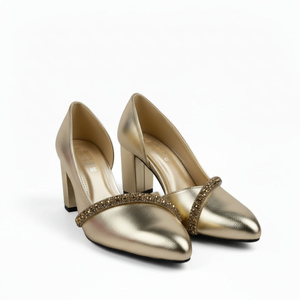 A pair of metallic gold pointed-toe high heels with a rhinestone-embellished cross-strap, displayed on a white studio floor.