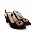 A pair of burgundy velvet slingback high heels with gold square buckle embellishments, displayed on a white studio floor.
