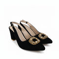 A pair of black pointed-toe slingback heels with chunky block heels and ornate gold-tone square embellishments on the toes, photographed on a white studio floor.