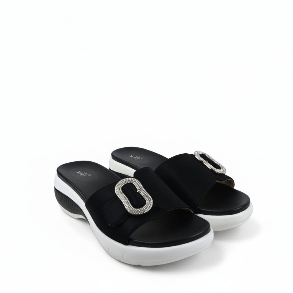 A pair of black platform slide sandals with a prominent silver-tone rectangular buckle embellished with clear rhinestones, shown on a white fabric backdrop.