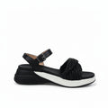 A black platform wedge sandal with a textured woven strap detail, shown from a side profile against a white fabric backdrop.