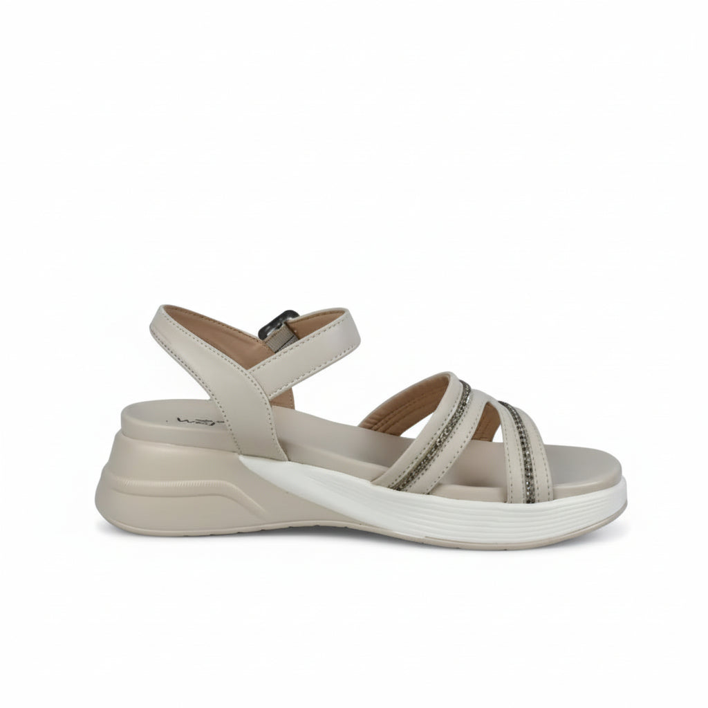 A single women's wedge sandal in a light cream or off-white color, featuring double straps with silver rhinestone embellishments and a low wedge heel, photographed against a wrinkled light gray background.