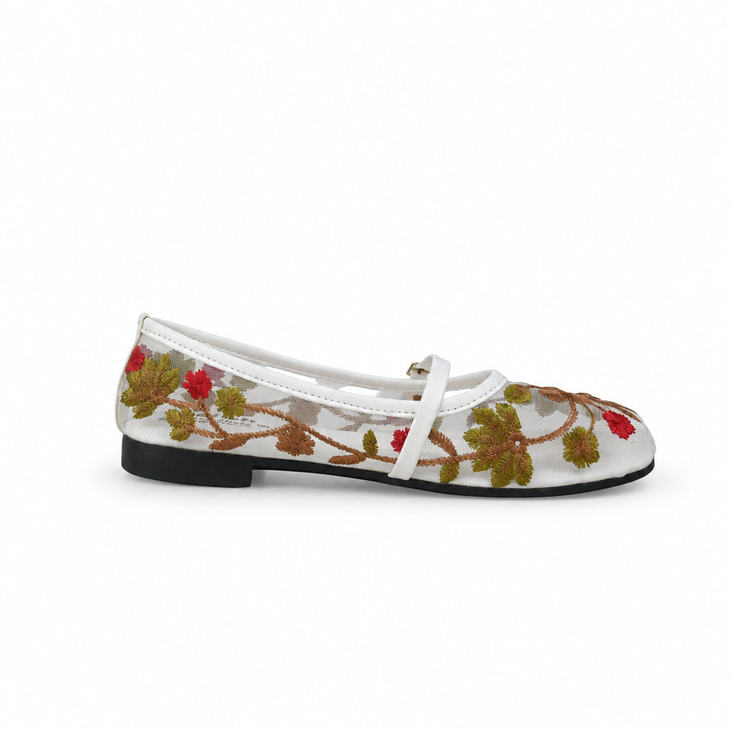 A white flat shoe with a colorful floral vine pattern in green, red, and yellow, featuring a white strap across the top and a black sole, shown against a wrinkled white backdrop.