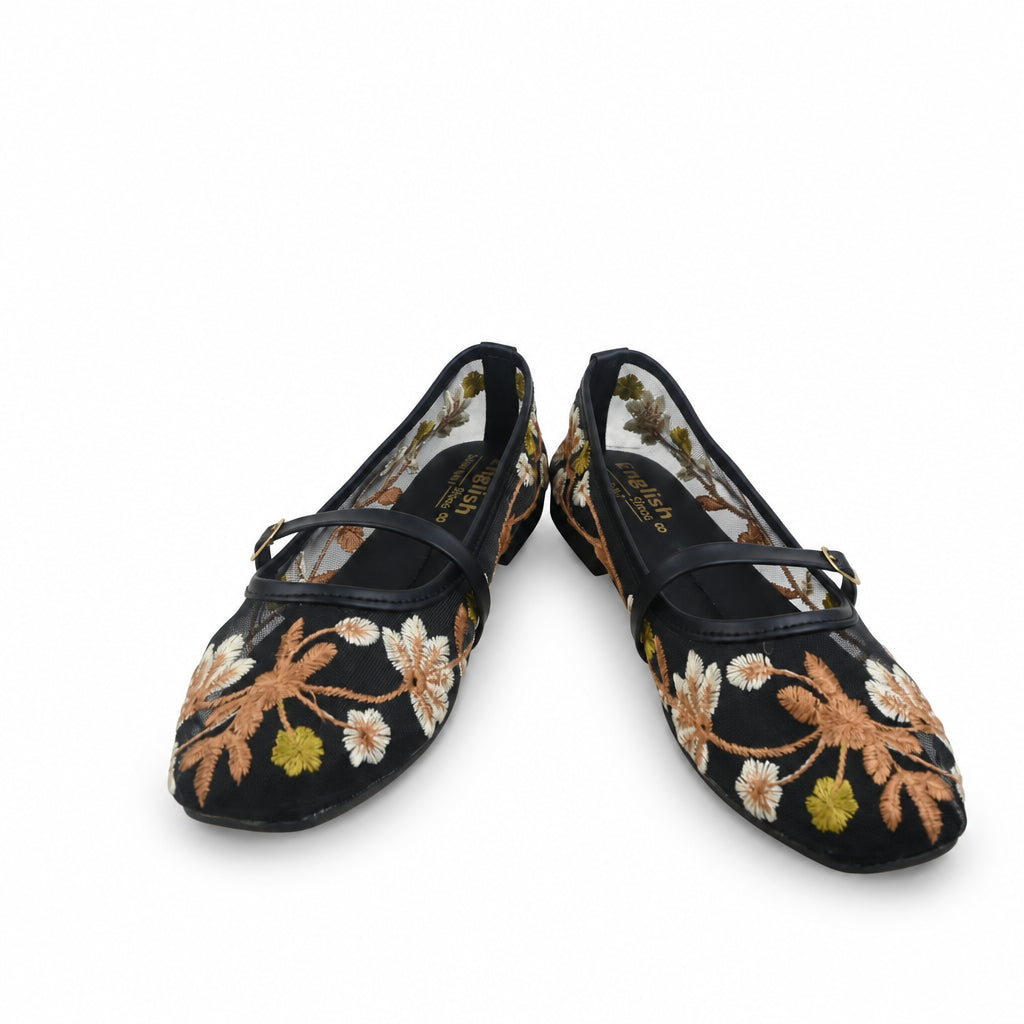 A pair of black Mary Jane flats with floral embroidery in gold, white, and green, shown next to their black and white packaging box on a white background.