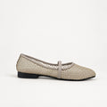 A single women's flat shoe with a woven beige upper and a thin beige strap across the instep, shown against a light gray backdrop. A partial black brand tag is visible on the right edge.