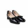 A pair of black patent leather block-heel pumps with bow details on the toes, displayed on a light gray textured surface.