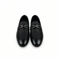 A pair of black loafers with a textured fabric upper and a shiny black leather strap across the vamp, displayed on a white surface next to an orange shoebox.