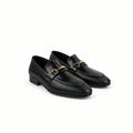 A pair of black leather loafers with a textured chevron pattern and a gold-tone horsebit detail across the vamp, displayed on a white fabric backdrop.