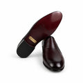A pair of men's leather dress shoes, with one shoe standing upright to show the dark brown polished upper and the other lying on its side to show the dark red-brown suede or leather outsole with a gold brand plaque. A blue shoebox is partially visible on the right against a white backdrop.