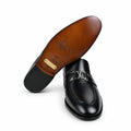 A pair of black leather penny loafers with a silver-tone LV monogram logo on the vamp, shown with one shoe turned to display the brown suede sole and the other standing upright. A blue and white Louis Vuitton shoebox is partially visible on the right.