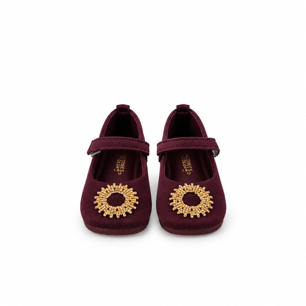 A pair of dark burgundy ballet flats with gold circular floral embellishments on the toes, displayed on a white surface with a white backdrop.