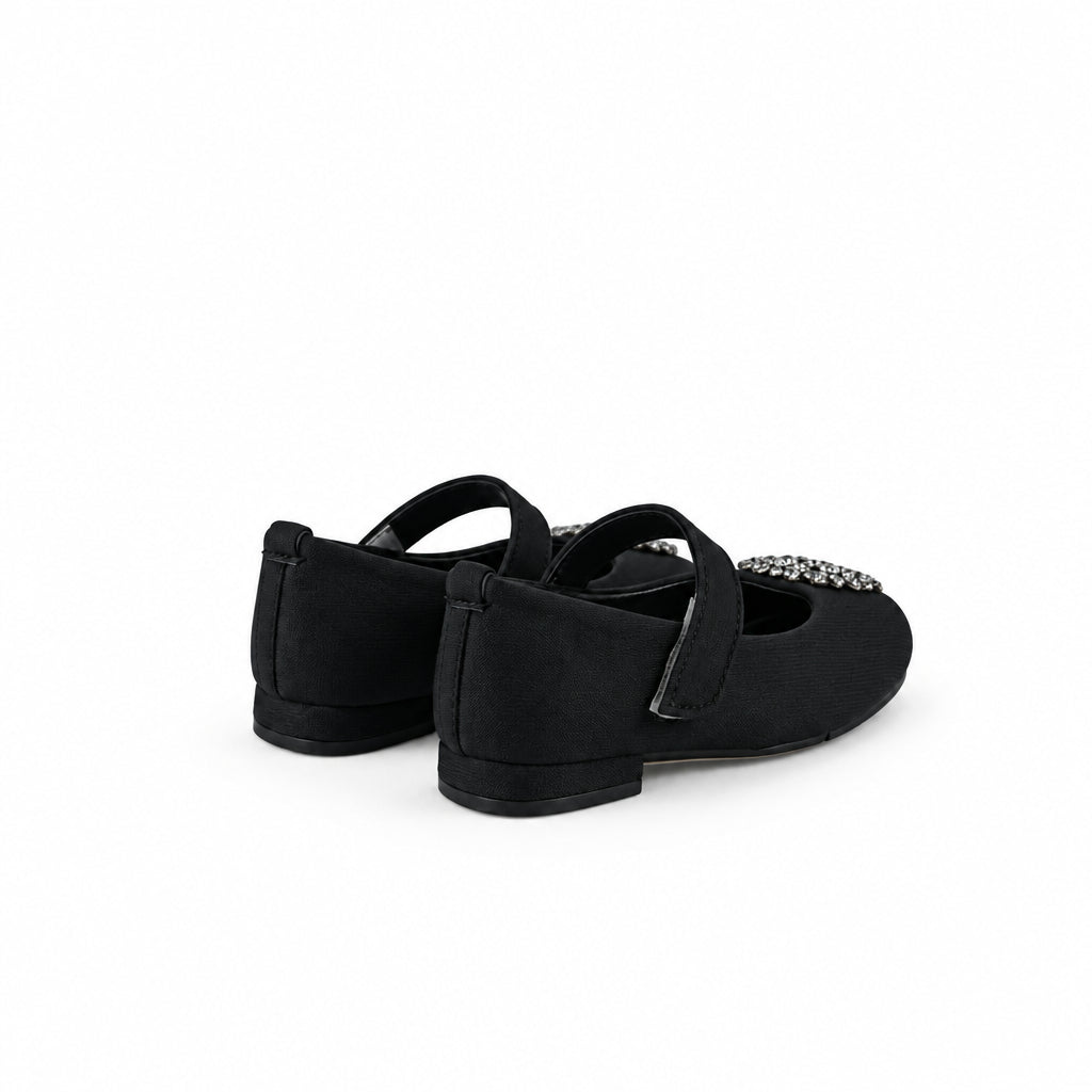 A pair of black flat shoes with decorative silver embellishments on the toe, photographed on a white fabric backdrop.