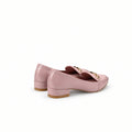 A pair of light pink patent leather loafers with a decorative bow featuring gold lettering, displayed on a white studio backdrop.